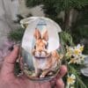 Uovo di Pasqua decorativo Blanc Mariclò con coniglio su sfondo blu, fiocco in raso e motivi floreali. Disponibile in due dimensioni.