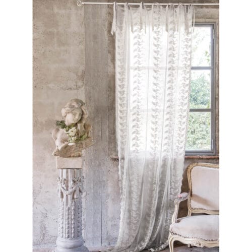 Tenda ricamata Blanc Mariclò in cotone panna, collezione Dentelle, appesa a una finestra accanto a una statua decorativa e una poltrona stile shabby chic.