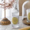 Bottiglia di profumo per tessuti Mathilde M. Mimosa Joli da 75 ml, con elegante design in vetro scanalato, accanto a una candela profumata sotto una campana di vetro e una decorazione shabby chic con dettagli floreali.
