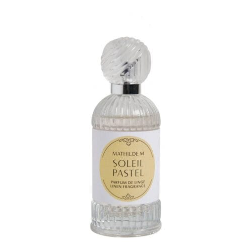 Bottiglia di profumo per tessuti Mathilde M. Soleil Pastel da 75 ml, accanto a una candela accesa, su sfondo decorativo con conchiglie e coralli in stile marino.
