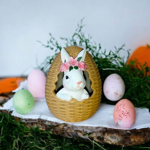 Coniglio decorativo con coroncina floreale che sbuca da un cesto a forma di uovo, perfetto per la Pasqua e l’arredamento primaverile.