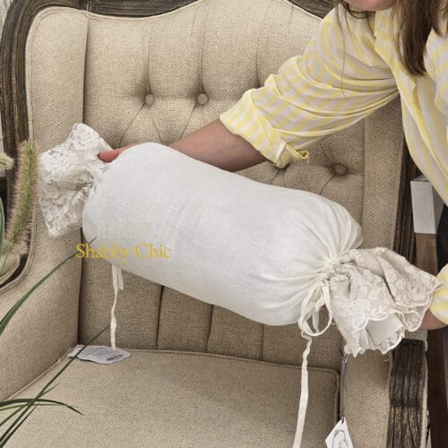 Fodera cuscino a forma di caramella in tessuto bianco con estremità arricciate in pizzo color fango, appoggiata su una poltrona imbottita beige.