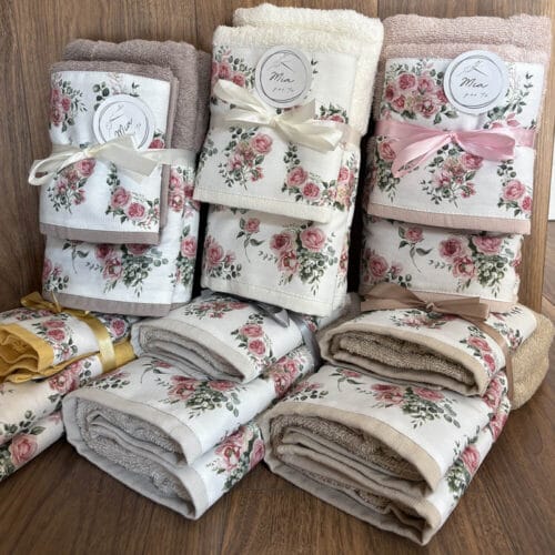 Set asciugamani in cotone viso e ospite con fascia decorativa floreale e fiocco, disponibili in sei colori pastello, disposti su fondo in legno.