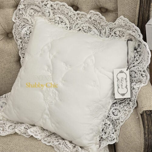 Fodera cuscino quadrata trapuntata in bianco naturale con balza in pizzo ricamato, appoggiata su poltrona imbottita in tessuto beige.