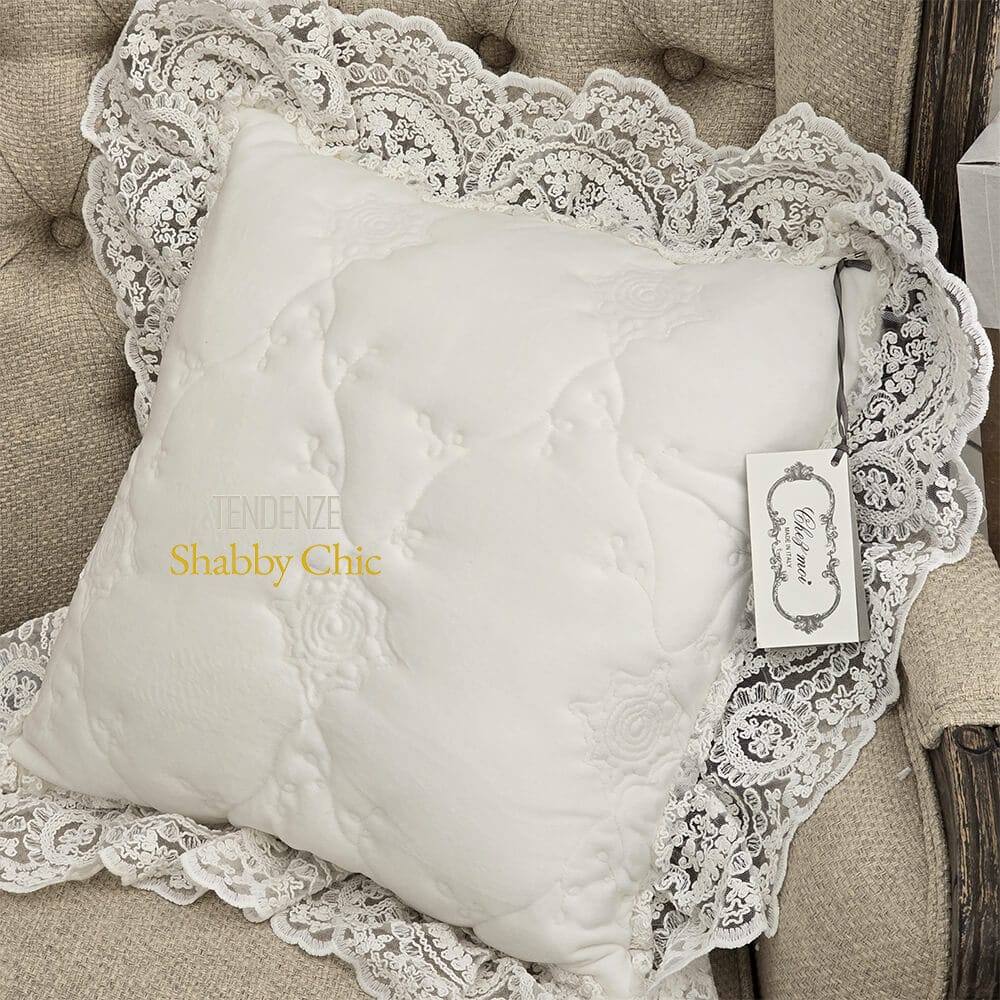 Fodera cuscino quadrata trapuntata in bianco naturale con balza in pizzo ricamato, appoggiata su poltrona imbottita in tessuto beige.
