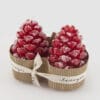 Due candele rosse a forma di pigna con effetto neve decorativo, confezionate con nastro e supporto in cartone.