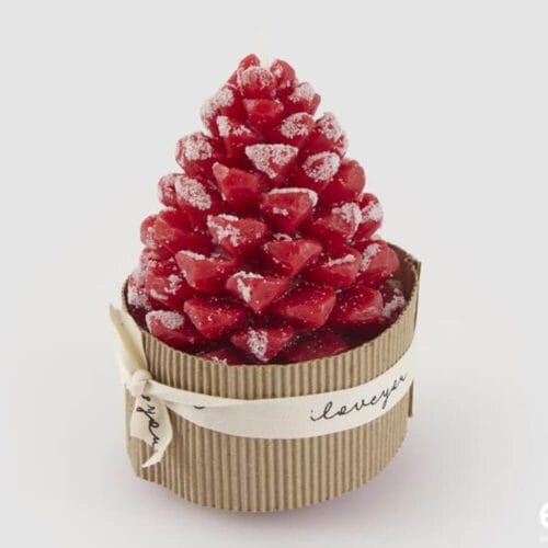 Candela rossa a forma di pigna innevata, confezionata con supporto in cartone e nastro decorativo.