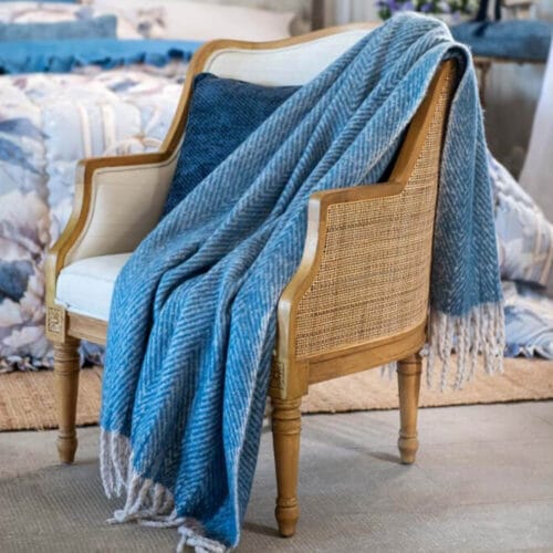 Plaid azzurro con motivo a spina di pesce e frange bianche, arrotolato e disposto elegantemente su una poltrona.