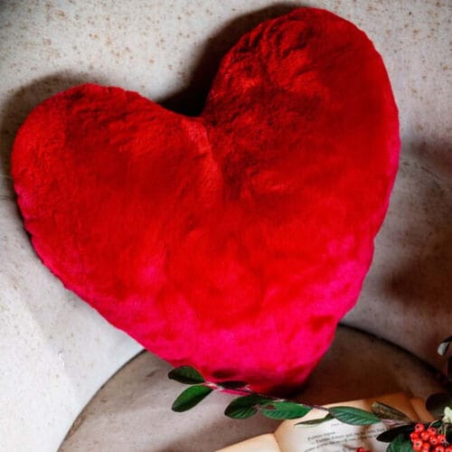 Cuscino a forma di cuore in ecopelliccia, disponibile nei colori beige e rosso, dalla texture morbida e avvolgente.