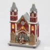 Église de Noël EDG avec murs rouges, toit gris enneigé, lumières LED et chanteurs de Noël sur le portail, décorations festives et fenêtres illuminées.