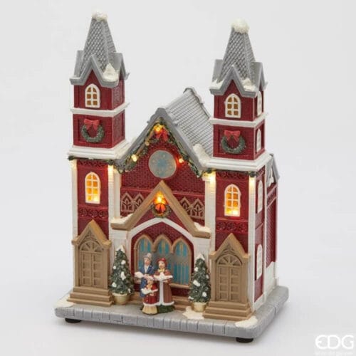 Chiesa natalizia EDG con pareti rosse, tetto grigio innevato, luci LED e gruppo di cantori sul portale, decorazioni festive e finestre illuminate.