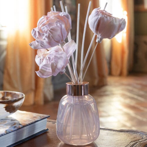 Diffusore per ambienti Mathilde M con fiori decorativi rosa in manioca e bastoncini in rattan, contenuto in un elegante flacone in vetro rosato iridescente.