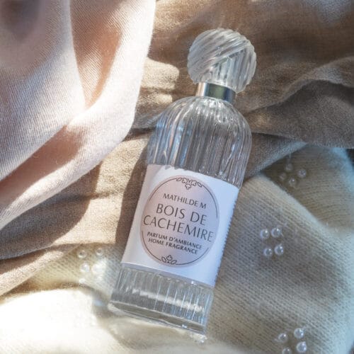 Flacone di profumo per tessuti Mathilde M Bois de Cachemire da 75 ml in vetro trasparente con scanalature verticali, adagiato su morbidi tessuti beige e crema.