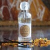 Flacone in vetro scanalato con tappo effetto cristallo contenente profumo per tessuti Mathilde M - Douceur d’Ambre, circondato da bastoncini di vaniglia e granuli ambrati.