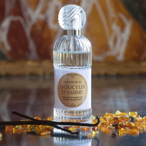 Flacone in vetro scanalato con tappo effetto cristallo contenente profumo per tessuti Mathilde M - Douceur d’Ambre, circondato da bastoncini di vaniglia e granuli ambrati.