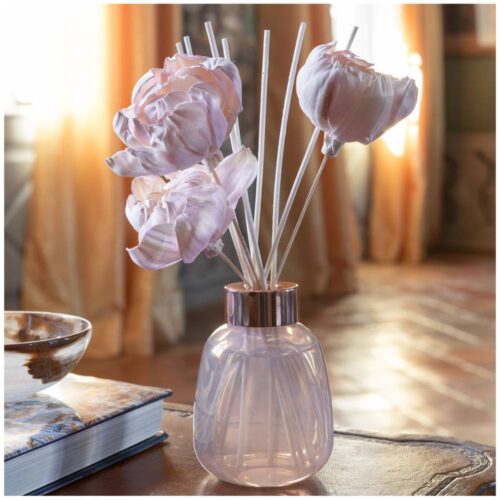 Diffusore con flacone in vetro iridescente, fiori rosa in manioca e bastoncini in rattan bianco, su sfondo elegante da interno.