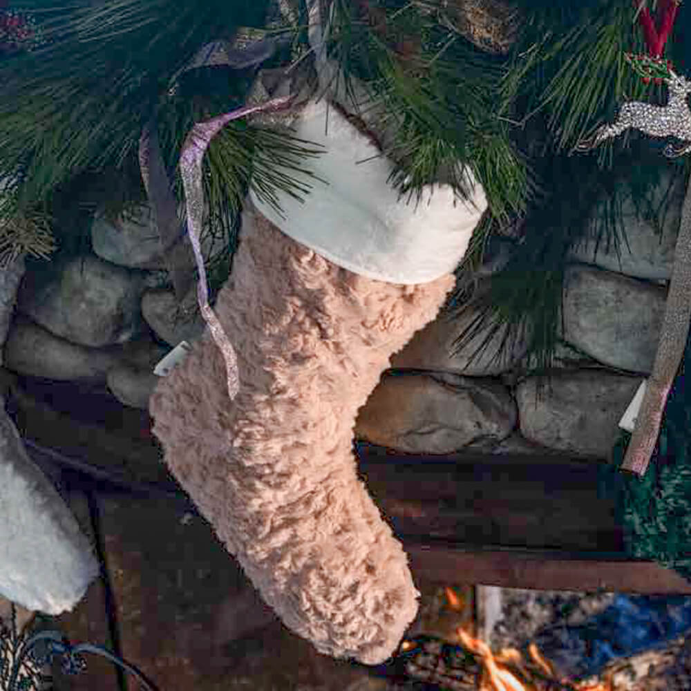 L’Atelier 17 Calza Pelliccia Let’s Do Xmas Rosa h 43 1 Calza natalizia rosa in pelliccia effetto bouclé con bordo bianco, appesa a un camino decorato con rami di pino.