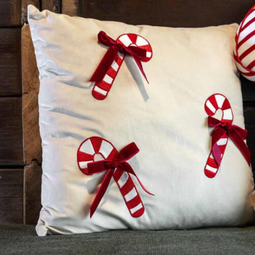 Cuscino natalizio 40x40 con ricami di candy cane rossi e bianchi e fiocchetti in velluto rosso, su fondo avorio.