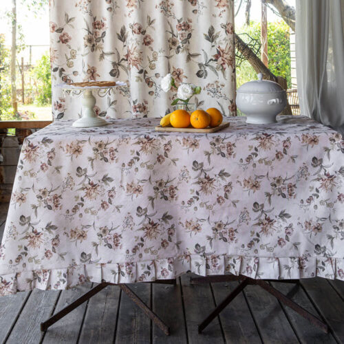 Tovaglia in cotone resinato color avorio con stampa floreale nei toni cipria e verde, bordata con balza arricciata coordinata.