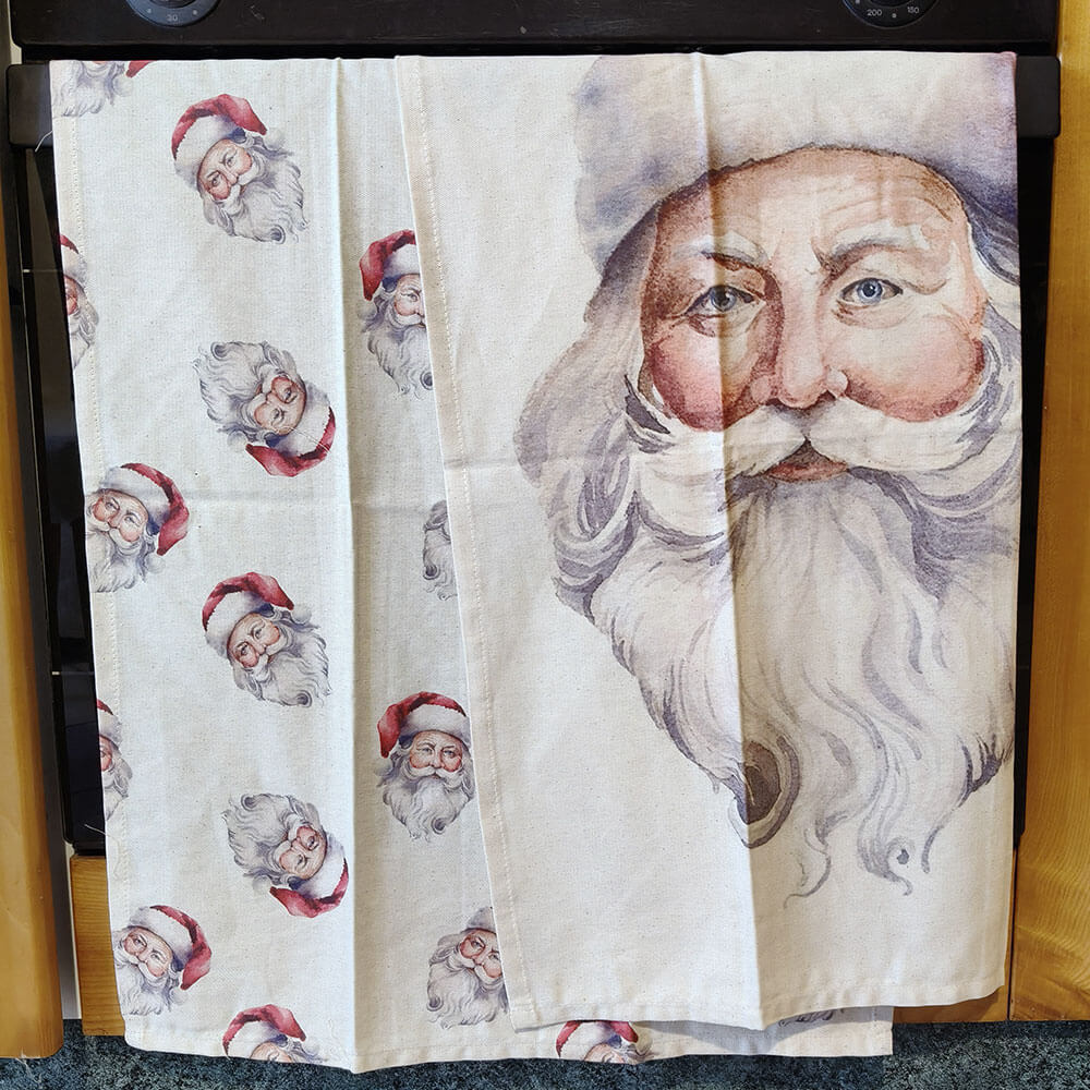 Lot de deux torchons de Noël en coton : l'un avec une impression répétée du visage du Père Noël, l'autre avec un portrait aquarelle en pied sur fond clair.