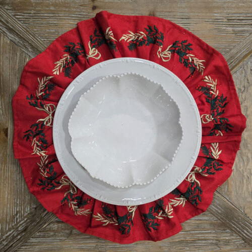 Tovaglietta natalizia rotonda in cotone rosso con balza plissettata decorata da ricami dorati, verdi e rossi a tema natalizio.