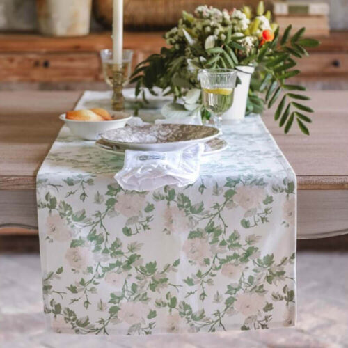 Chemin de table Blanc MariClò avec un motif floral de roses pâles et de brins verts sur une base crème, sur un set de table en bois.