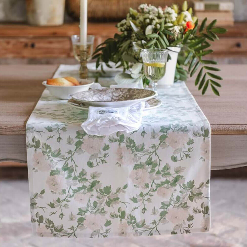 Chemin de table Blanc MariClò avec un motif floral de roses pâles et de brins verts sur une base crème, sur un set de table en bois.