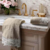 Set di asciugamani beige con bordo in pizzo bianco floreale, posati su lavabo in marmo con dettagli in ottone.