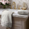 Set asciugamani color panna con bordo in pizzo floreale bianco, disposti su lavabo in marmo con rubinetteria dorata.