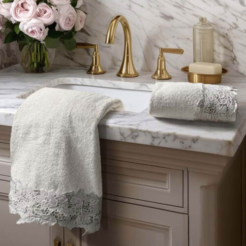 Set asciugamani color panna con bordo in pizzo floreale bianco, disposti su lavabo in marmo con rubinetteria dorata.