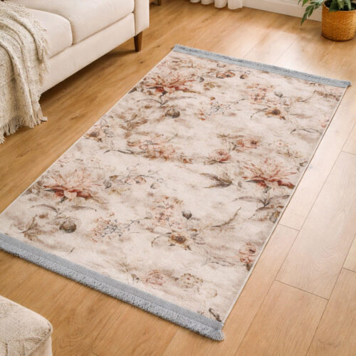Tappeto in velluto beige con stampa floreale nei toni caldi e frange grigio perla, posato su pavimento in legno chiaro.