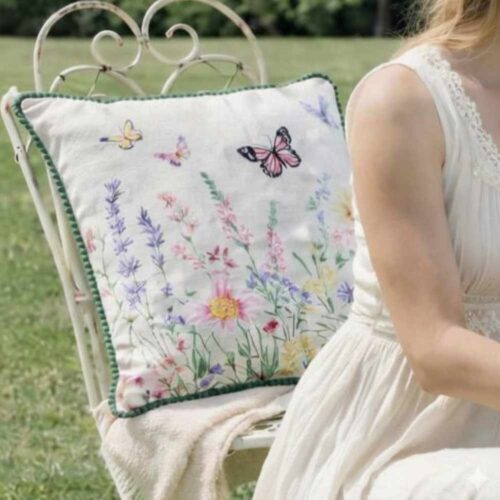 Cojín de mobiliario crema serie Clodette Lucilla con bordado de flores campestres lilas y amarillas sobre tejido texturizado efecto lino.