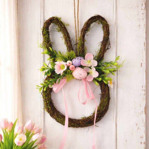 Ghirlanda a forma di coniglio in vimini con fiori rosa, foglie verdi, ovetti decorativi e fiocco rosa.
