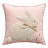 Coussin carré rose poudré avec application de lapin en tissu bouclé, nœud de cou beige et petite broderie florale.