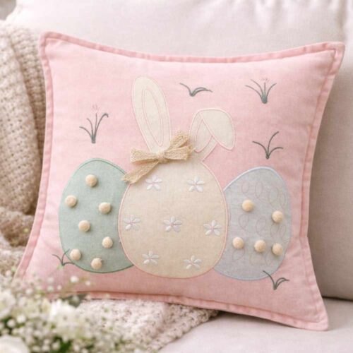 Cuscino rosa cipria con applicazioni a uova pastello, coniglietto stilizzato con fiocco beige e pom pon decorativi.