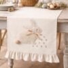 Camino de mesa crema con bordado de conejo en tejido bouclé, lazo beige y pequeñas flores bordadas, con volante final.