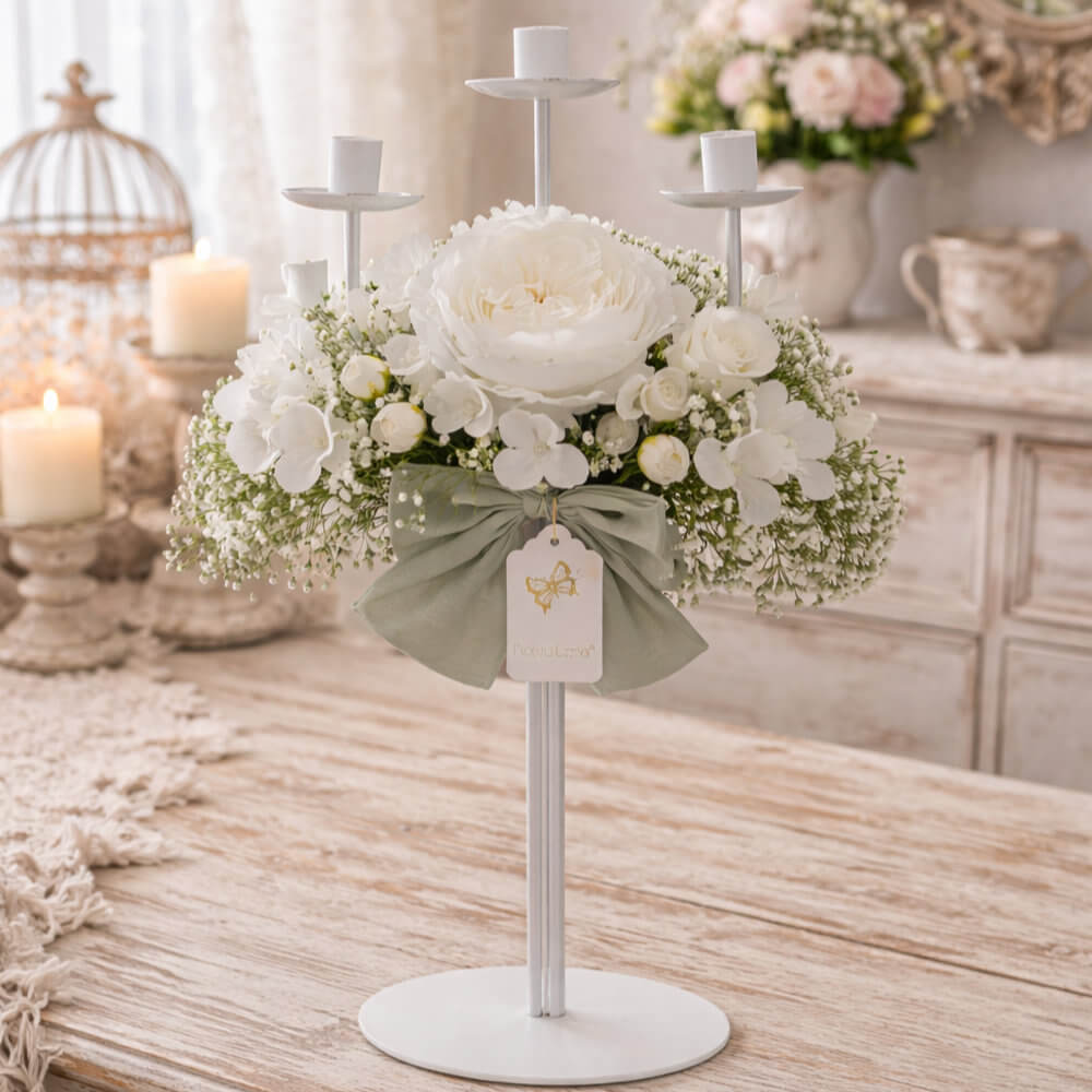 Candelero Fiori di Lena de 3 llamas con dos peonías blancas y adornos florales blancos, lazo verde salvia, sobre una mesa de madera Shabby Chic.