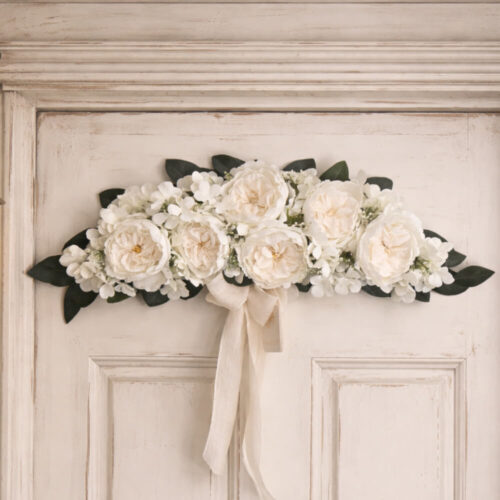 Colgante de pared floral Fiori di Lena con 6 peonías blancas, hortensias y muérdago, lazo claro y vegetación decorativa.