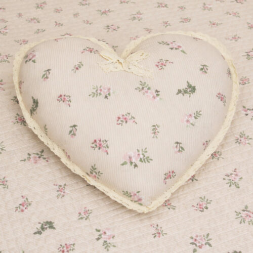 Cuscino decorativo a forma di cuore Fiore di Primavera L'Atelier 17 in poliestere avorio a righe con stampa floreale rosa, bordo in pizzo all'uncinetto e fiocchetto decorativo, posato su trapuntino coordinato.