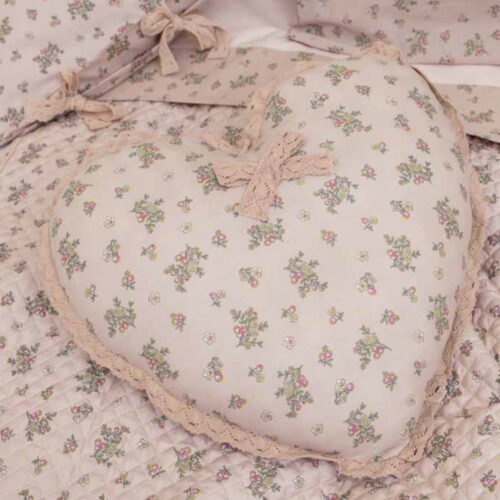 Cuscino decorativo a forma di cuore Cuor di Magnolia L'Atelier 17 in poliestere rosa cipria con stampa floreale multicolore, bordo in pizzo all'uncinetto e laccetto decorativo in pizzo annodato, posato su trapuntino coordinato.