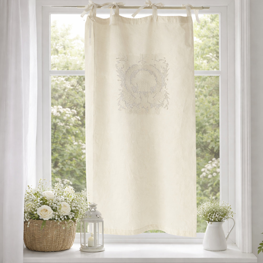 Cortina Sissi Avorio L'Atelier 17 de algodón marfil con bordado central tono sobre tono de un medallón heráldico con volutas y motivos barrocos, sujeta con presillas con lazos, instalada en ventana de estilo Shabby Chic.