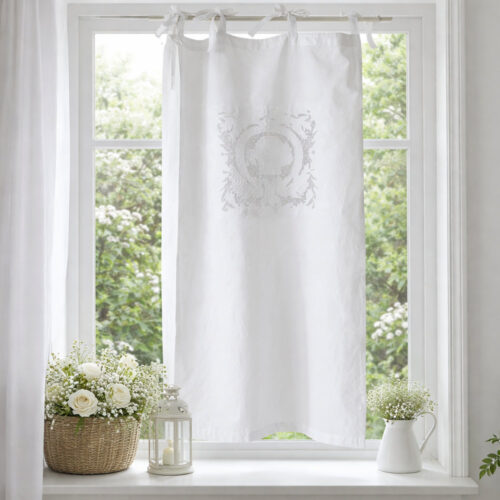Cortina Sissi L'Atelier 17 de algodón blanco con bordado central tono sobre tono de un medallón heráldico con volutas y motivos florales barrocos, sujeta con lazos de raso blanco.
