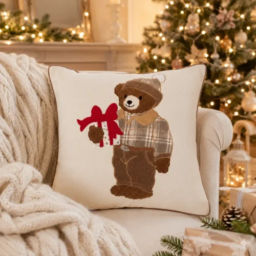 Cuscino Blanc Mariclò color avorio con applicazione di un orsetto in tessuto effetto teddy, abbigliato con cappello e abiti tartan beige, mentre tiene un pacco regalo con grande fiocco rosso.