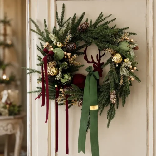 Ghirlanda Guardiano del Bosco seduto 50 cm con renna bordeaux, sfere verdi e oro, pigne, abete e lunghi nastri.