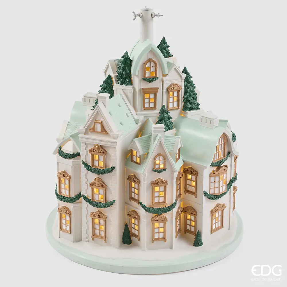Base per albero EDG a forma di palazzi bianchi e verde acqua, con finestre dorate illuminate, abeti e ghirlande verdi.