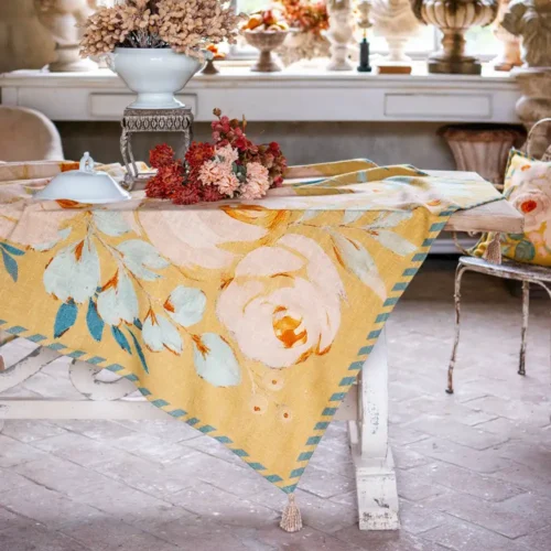 Copritutto Bloom Collection color ocra con rose chiare, foglie verde salvia, bordo a righe e nappine decorative.
